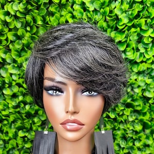 May include: A mannequin head with short, gray hair styled in a side-swept pixie cut. The mannequin has full makeup, including long eyelashes and dark gray fringed earrings. The background is a wall of green foliage.