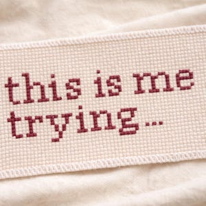 Puede incluir: Un parche rectangular de punto de cruz con la frase "this is me trying..." en hilo burdeos. El parche está hecho de una tela color crema con un borde cosido, sobre una superficie suave y blanquecina.
