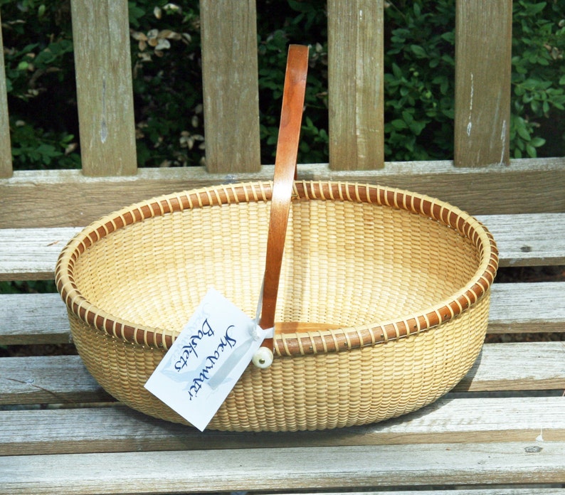 May include: A woven basket with a brown leather handle. The basket is oval shaped and has a light brown color. A white tag with blue text reads "Greenwater Baskets".