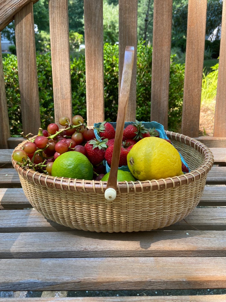 May include: A woven basket with a wooden handle filled with red grapes, strawberries, limes, and a lemon. The basket is sitting on a wooden bench.