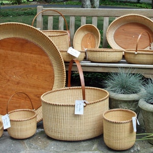 May include: A collection of woven baskets of various sizes and shapes, including a large round tray with a wooden insert, a tall basket with a leather handle, and several smaller baskets. The baskets are arranged on a wooden bench and on the ground, with some potted plants in the background.