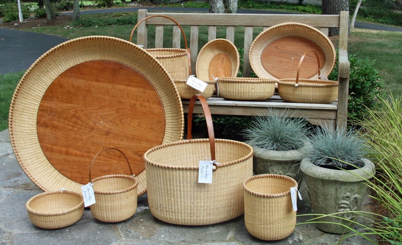 May include: A collection of woven baskets of various sizes and shapes, including a large round tray with a wooden insert, a tall basket with a leather handle, and several smaller baskets. The baskets are arranged on a wooden bench and on the ground, with some potted plants in the background.