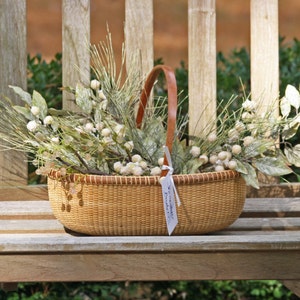 May include: A woven basket filled with white berry sprigs and a brown leather handle. The basket is sitting on a wooden bench.