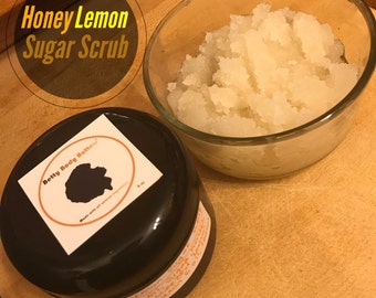 May include: A close-up of a glass bowl filled with a white sugar scrub. The bowl is sitting on a wooden surface. The text "Honey Lemon Sugar Scrub" is visible in the background. The label on the black jar reads "Betty Body Butter".