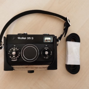 May include: A black Rollei 35S camera with a leather strap and a black leather case. The camera has a silver dial and a silver lens.
