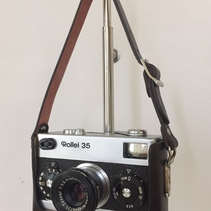 May include: A silver Rollei 35 camera with a brown leather case and a black strap. The camera has a lens labeled "Tessar 1:3.5 f=40mm" and a dial for adjusting the color setting.