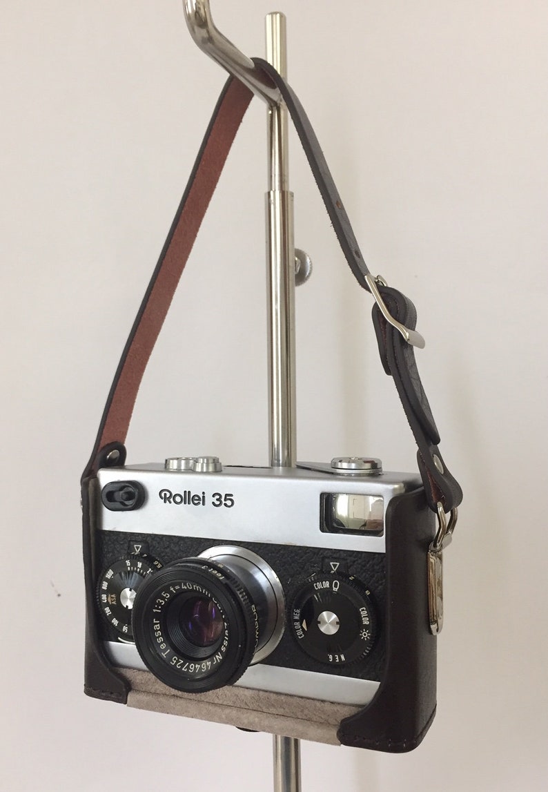 May include: A silver Rollei 35 camera with a brown leather case and a black strap. The camera has a lens labeled "Tessar 1:3.5 f=40mm" and a dial for adjusting the color setting.