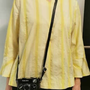 May include: A black and silver camera with a black leather strap hanging around a person's neck. The camera has the text "Goliel 35 S" on the front. The person is wearing a yellow and white striped shirt.