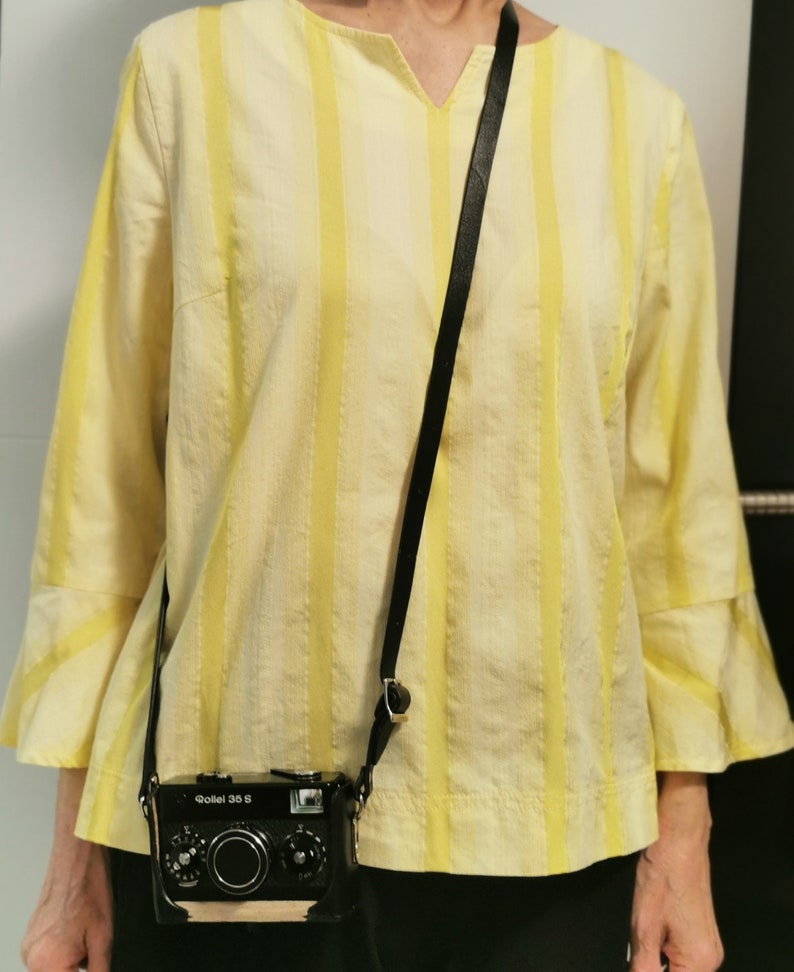 May include: A black and silver camera with a black leather strap hanging around a person's neck. The camera has the text "Goliel 35 S" on the front. The person is wearing a yellow and white striped shirt.