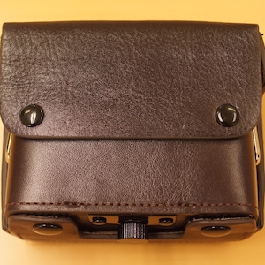 May include: A brown leather camera case with a flap closure and metal accents. The case has two metal snaps on the front and a metal buckle on the side.