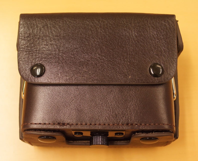 May include: A brown leather camera case with a flap closure and metal accents. The case has two metal snaps on the front and a metal buckle on the side.