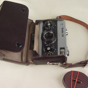 May include: A silver Rollei 35 camera with a brown leather case and strap. The camera has a black lens and a silver body.