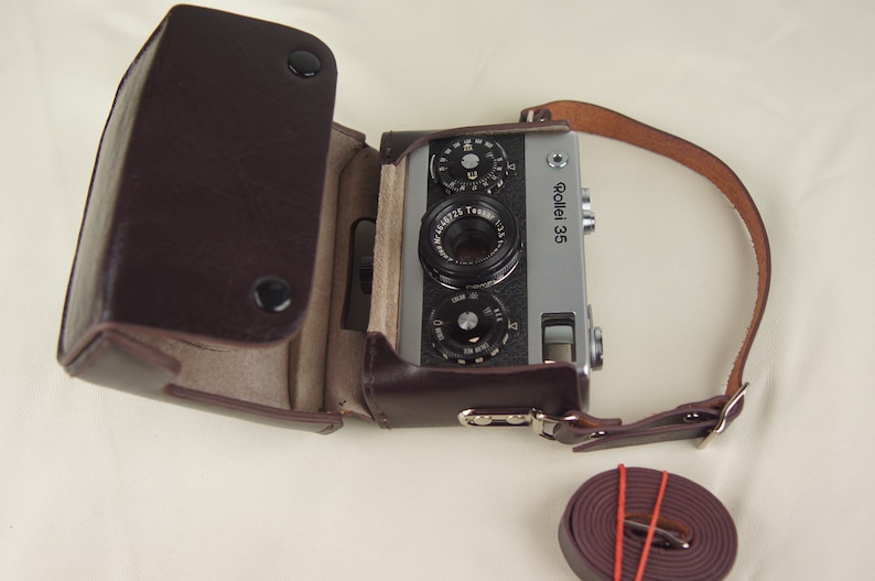 May include: A silver Rollei 35 camera with a brown leather case and strap. The camera has a black lens and a silver body.