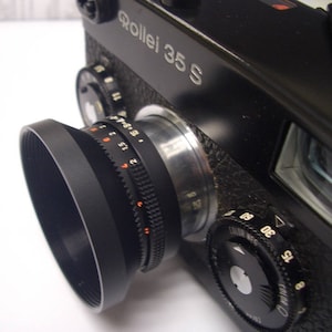 May include: A black Rollei 35S camera with a lens hood and a silver lens ring. The camera has a black leatherette covering and a silver nameplate that reads "Rollei 35 S". The lens has a depth of field scale and a focus ring. The camera has a shutter speed dial and a film speed dial.