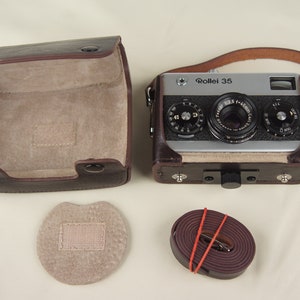 May include: A brown leather camera case with a matching strap, a brown leather camera case lid, and a brown leather camera strap. The camera case is open and the camera is visible inside. The camera is a silver Rollei 35 with a black lens. The camera has a black leather strap attached to it.
