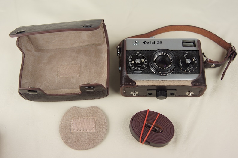 May include: A brown leather camera case with a matching strap, a brown leather camera case lid, and a brown leather camera strap. The camera case is open and the camera is visible inside. The camera is a silver Rollei 35 with a black lens. The camera has a black leather strap attached to it.