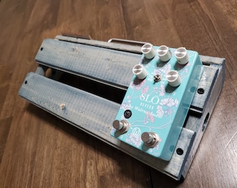 May include: A turquoise blue guitar effects pedal with a floral design. The pedal is labeled "SLO REVERB by Walrus Audio" and has five knobs. The pedal is sitting on a blue wooden pedalboard.