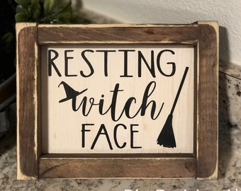 May include: A wooden sign with a light background and a dark brown frame. The sign reads "RESTING witch FACE" in black lettering, with a witch hat and broom graphic. The sign is a Halloween-themed decoration.