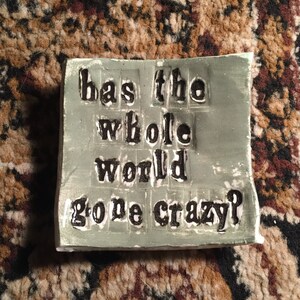May include: A green ceramic tile with the words "has the whole world gone crazy?" printed in black.