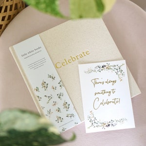 以下が含まれることがあります： 「Celebrate」のゴールド文字が入ったクリーム色のジャーナルとカードのセット。カードには「There's always something to Celebrate!」のテキストと花のアクセントがあります。「little white books」のテキストが入ったブックマークも表示されています。
