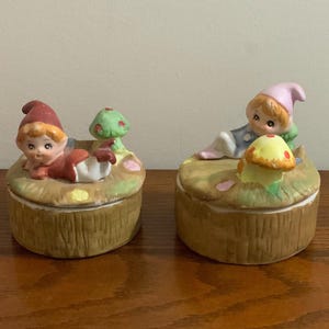 Puede incluir: Dos cajas de cerámica para guardar pequeños objetos con un tema de setas y duendes. Una caja tiene un duende con un gorro rojo tumbado junto a una seta verde. La otra caja tiene un duende con un gorro rosa sentado junto a una seta amarilla.