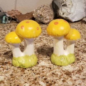 Puede incluir: Un juego de dos saleros y pimenteros de cerámica con forma de hongo. Los hongos son amarillos con puntos naranjas y tienen tallos blancos y bases verdes.