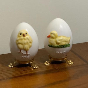 Puede incluir: Dos huevos de cerámica blanca con soportes dorados. Un huevo presenta un pollito amarillo con ojos negros y pico rojo. El otro huevo presenta un pato amarillo con pico rojo y patas naranjas. Ambos huevos tienen el año 1972 impreso.