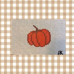 May include: A needlepoint canvas featuring a centered orange pumpkin design with a brown stem, worked on a white grid mesh. The bottom right corner displays the black initials BK stitched into the fabric, creating a clear, geometric pattern.