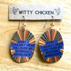 Könnte beinhalten: Ohrringe in ovaler Form mit einem farbenfrohen, abstrakten Design in Orange, Braun und Blau. Jeder Ohrring hat ein blaues Rechteck mit dem Text "SORRY I COULDN'T HEAR YOU OVER MY INTERNAL MONOLOGUE." Die Ohrringe sind von "WITTY CHICKEN."