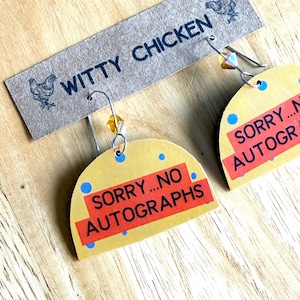 Könnte beinhalten: Handgefertigte Ohrringe mit gelbem, halbkreisförmigem Design. Die Ohrringe tragen den Text "SORRY...NO AUTOGRAPHS" auf einem roten Rechteck. Die Ohrringe sind an einem Anhänger mit der Aufschrift "WITTY CHICKEN" mit einer Hühnerillustration befestigt.