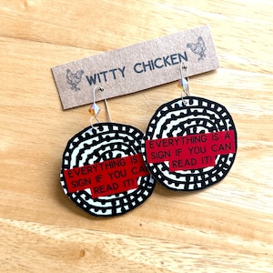 May include: Black and white circular earrings with a spiral design and a red banner that reads "EVERYTHING IS A SIGN IF YOU CAN READ IT!". The earrings are from "WITTY CHICKEN".