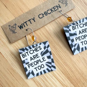 Könnte beinhalten: Quadratische Ohrringe mit dem Text "BITCHES ARE PEOPLE TOO" in Schwarz und Weiß, vor einem karierten Hintergrund. Die Ohrringe sind an silbernen Haken mit einer kleinen orangefarbenen Perle befestigt. Ein braunes Etikett oben zeigt "WITTY CHICKEN" mit Hühnerillustrationen.