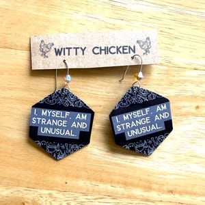 Könnte beinhalten: Sechseckige Ohrringe mit dem Text "I, MYSELF, AM STRANGE AND UNUSUAL" auf einem schwarz-weiß gemusterten Hintergrund. Die Ohrringe sind an einem braunen Etikett mit den Worten "WITTY CHICKEN" und Hühnerillustrationen befestigt.
