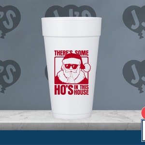 May include: White foam cup with a red graphic of Santa Claus wearing sunglasses and a finger to his lips. The text reads "There's some HO'S in this house". The cup is sitting on a white countertop with a blue background. The background also has a logo of three red hearts with the text "JJ's Party House".