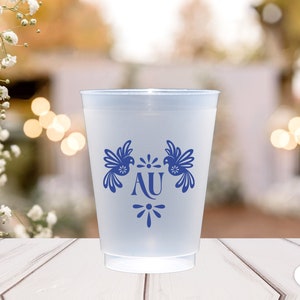 May include: A white plastic cup with a blue design of two birds and the letters "AU" in the center. The cup is on a white wooden surface.