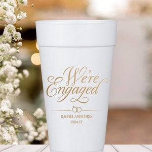 May include: White foam cup with the text "We're Engaged" in gold script. Below the text are two gold rings and the names "Rachel and Chris" with the date "03.04.2X".
