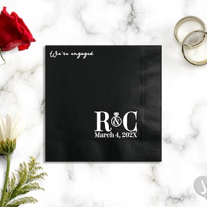 May include: Black paper napkin with white text that says "We're engaged" and "R&C" with a diamond ring symbol and the date "March 4, 202X".