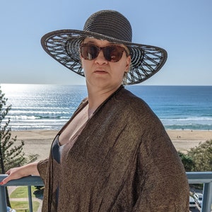 May include: A woman wearing a wide-brimmed black sun hat with a gold and black sheer cover-up over a black swimsuit. She is standing on a balcony overlooking the ocean.
