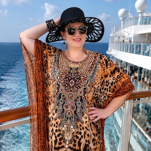 Könnte beinhalten: Ein locker sitzendes Kaftan-Kleid mit einem lebendigen Leopardenmuster, einem dekorativen, mit bunten Steinen und Perlen verzierten Ausschnitt und einem geometrischen Bordürenmuster am Saum. Das Kleid hat weite Ärmel und eine fließende Silhouette.