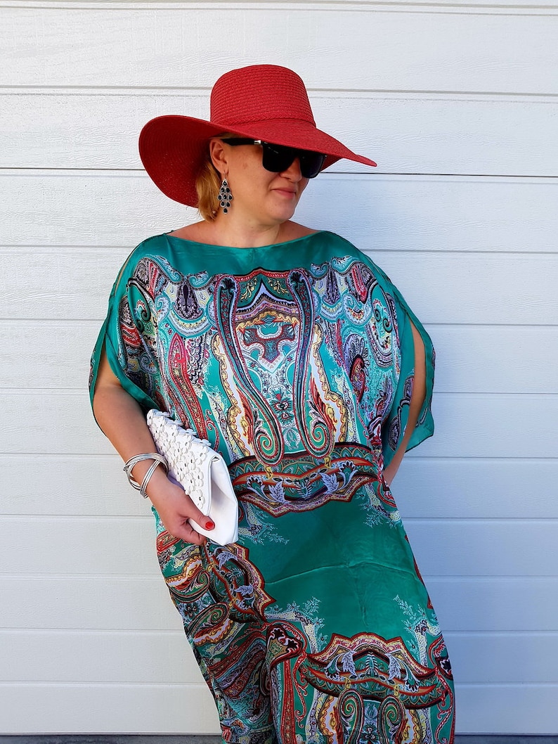 A woman wearing a green and blue paisley patterned maxi dress with a red wide-brimmed hat and a white clutch.
