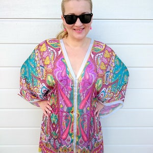May include: A woman wearing a pink, green, and blue paisley patterned kaftan with a V-neck and long sleeves. She is wearing sunglasses and pink earrings.