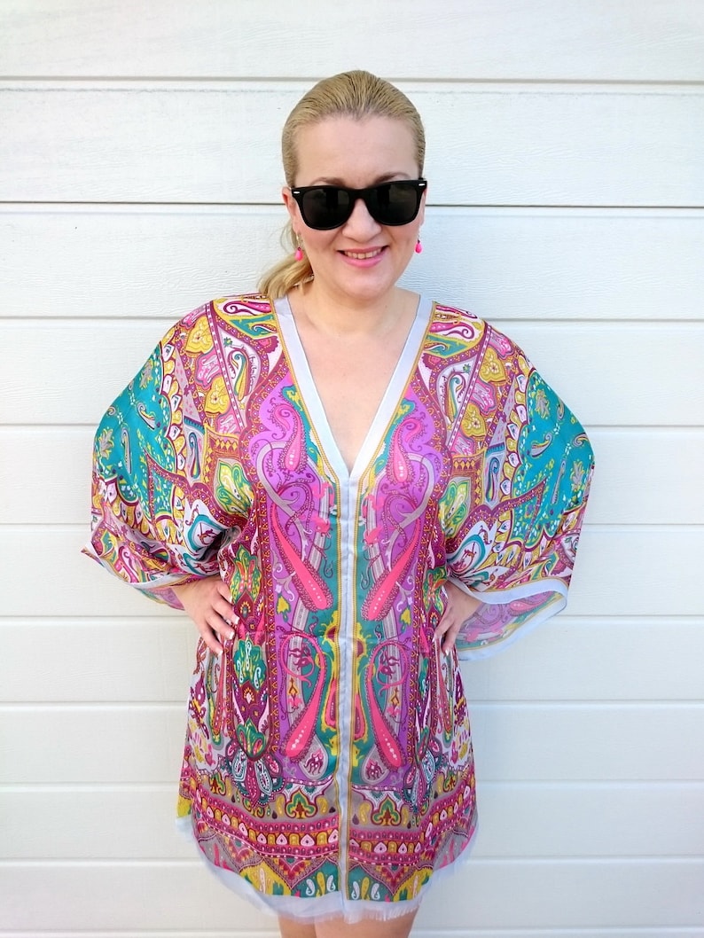 May include: A woman wearing a pink, green, and blue paisley patterned kaftan with a V-neck and long sleeves. She is wearing sunglasses and pink earrings.