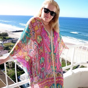 May include: A woman wearing a colorful patterned kaftan with a white background. The kaftan has a long, flowing design and features a variety of colors, including pink, blue, green, and yellow. The woman is standing on a balcony overlooking a beach.