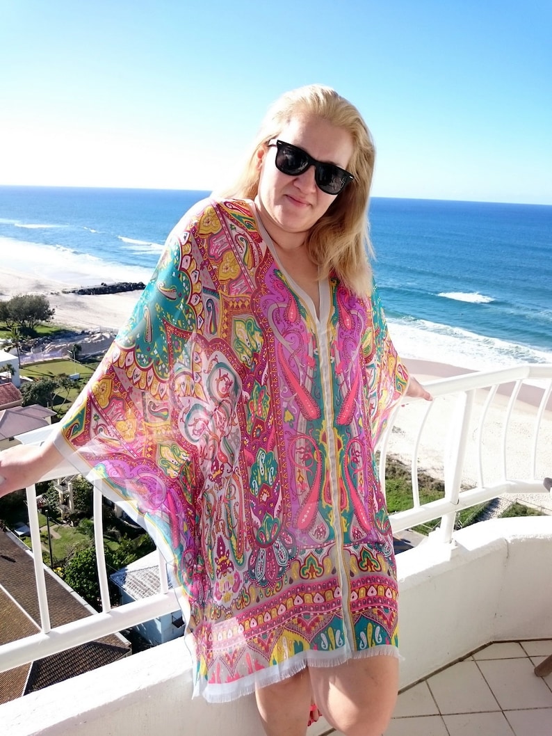 May include: A woman wearing a colorful patterned kaftan with a white background. The kaftan has a long, flowing design and features a variety of colors, including pink, blue, green, and yellow. The woman is standing on a balcony overlooking a beach.