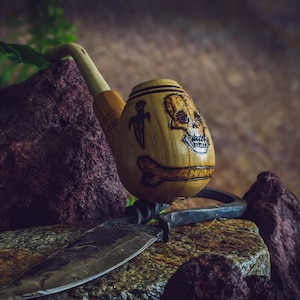 Könnte beinhalten: Eine Holzpfeife mit einem Totenkopfdesign und einem Knochenmotiv. Die Pfeife liegt auf einem Stein mit einem Messer aus Metall.