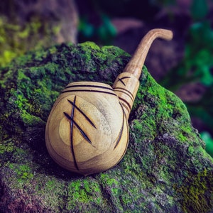 Puede incluir: Una pipa de madera con una cazoleta tallada y una boquilla larga. La cazoleta está decorada con un diseño tallado. La pipa está apoyada en una roca cubierta de musgo.