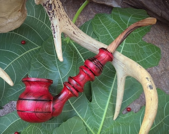 May include: A handcrafted, red wooden pipe with a curved stem, accented with black lines. The pipe rests on a bed of green leaves, with deer antlers and small red berries in the background. A unique, decorative smoking accessory.