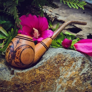 May include: A wooden pipe with a carved design of a star and crescent moons. The pipe is resting on a rock with pink flowers around it.