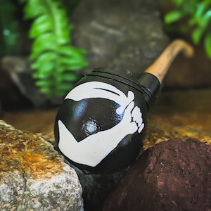 May include: A black wooden pipe with a white pirate symbol on the bowl. The symbol is a hand holding a sword.