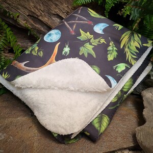 May include: A soft, black and white fleece blanket with a dark green, black, and white print of moons, antlers, leaves, and a pentagram. The blanket is folded in half and lying on a rock.